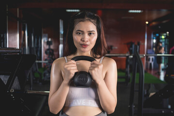 A young asian woman prepares to do a set of goblet squats with a kettlebell. Holding the weight in front of her chest. Training at a modern gym.