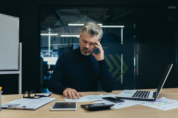 Serious thinking financier business analyst working with documents inside office, mature adult accountant on paper work calculating with calculator and laptop inside office.