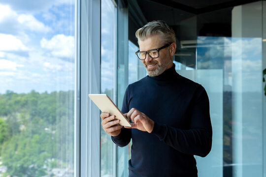 Mature adult businessman standing near window inside office, man smiling and using tablet computer, gray-haired boss browsing internet pages and reading news.