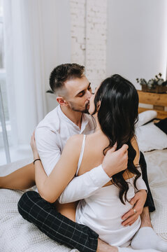 A stylish young bearded groom and a beautiful smiling brunette bride with a bouquet hug, kiss while sitting on the bed in the interior, indoors. Wedding photography of happy newlyweds, portrait.
