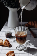 Transparent glass with black espresso coffee and pouring milk on a table set with croissants and jam, morning, breakfast, coffee break closeup concept.