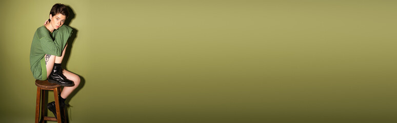 full length of trendy woman in black rough boots and striped pullover sitting on wooden stool on...