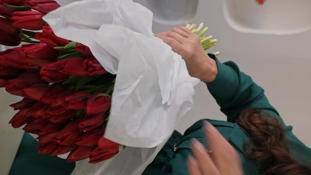 Caucasian lady florist collects a lush bouquet of tulips. A professional makes a fresh bouquet. Small business. A woman crumples paper (Crepe) and wraps a bouquet of tulips in it. Vertical video.