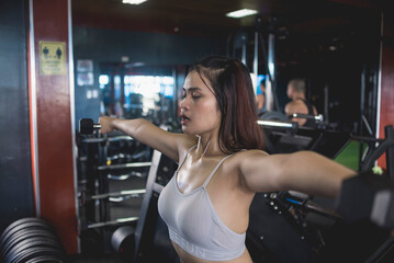 Fototapeta na wymiar An exhausted young woman powers through the last reps of side lateral dumbbells raises. Training and toning shoulder muscles at the gym.
