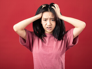 Fototapeta premium A portrait of a young Indonesian (Asian) woman wearing a pink shirt, looking dizzy and holding her head with both hands. Isolated with a red background