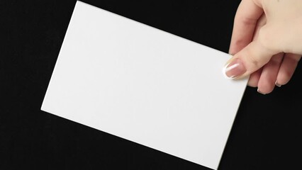 Vertical video. White card. Empty note. Invitation letter. Female hand showing clean mockup paper sheet with copy space on black background loop. - Powered by Adobe