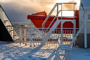 Part of orange lifeboat on snow and ice-covered ferry deck, dramatic and sunny sky in sunset. Norway.