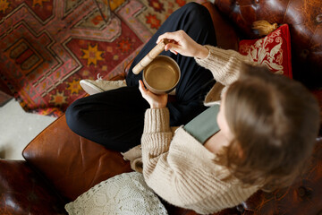 Young pretty caucasian girl playing on chakra cleaning tibetan singing bowl, while sitting on sofa...