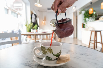 Waiter pouring red tea in a cup by a cast iron teapot. Man serving hot drinks in a cafeteria.