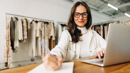 Portrait of a clothing store owner writing in a notebook and using a laptop