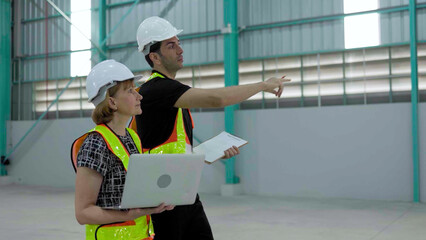 Caucasian senior female manager wearing safety vest holding laptop and male engineer walking warehouse inspection
