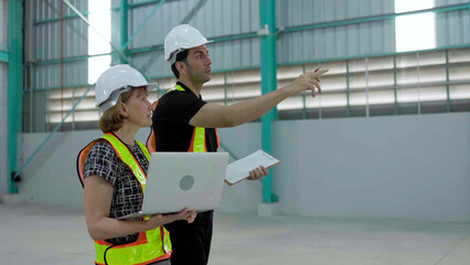 Caucasian senior female manager wearing safety vest holding laptop and male engineer walking warehouse inspection