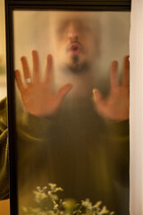 Men's portrait through a matte glass door at home. Weird man touching glass surface