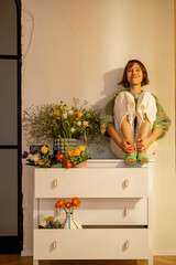 Young woman sits alone on white commode decorated with bouquets of flowers at home. Concept of beauty and fresh flowers indoors