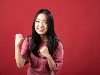 A studio portrait of a young Indonesian woman (Asian) wearing a pink shirt, seen celebrating and raising both hands. Isolated with a red background