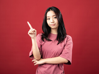 A studio portrait of a young Indonesian (Asian) woman wearing a pink shirt, seen pointing with one hand. Isolated with a red background