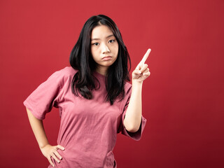 A studio portrait of a young Indonesian (Asian) woman wearing a pink shirt, seen pointing with one hand. Isolated with a red background