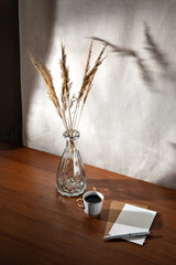 Aesthetic summer interior still life with cup of coffee, paper cards and dry grass in glass vase on wooden table, floral sunlight shadows on a wall, morning business concept