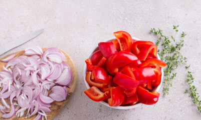 A bowl of chopped red sweet peppers, a wooden board with chopped red onions, fresh thyme sprigs and a kitchen knife on a beige textured background, top view