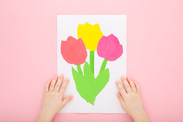 Little child hands and colorful tulip flowers with green leaves on white paper on light pink table...