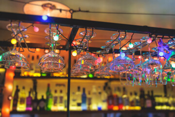 Bar interior details, glasses for wine and different alcohol cocktail drinks hang over bar counter with alcohol bottles and bartender in the background, nightlife club with backlit illumination
