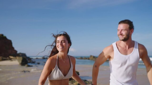 Young happy couple runs holding hands on beach through waves splashing water and enjoying summer closeup shot. Boyfriend and girlfriend having fun at seaside in spectacular scenic tropical location.