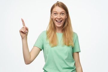 Indoor studio portrait of young ginger female with freckles point with a finger aside at copy space isolated over background