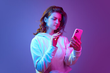 Young girl typing messages on mobile phone, reading news and social media against gradient blue purple studio background in neon light. Concept of emotions, youth, lifestyle, modern technologies