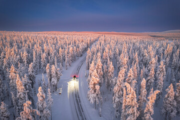 Aerial view of car on icy road and illuminated headlamps driving in the snowcapped forest, at dawn, Akaslompolo, Kolari, Pallas-Yllastunturi National Park, Lapland region, Finland