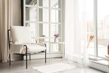 A white faux leather armchair near the window next to a small table with flowers. Part of the interior with copy space.High quality photo