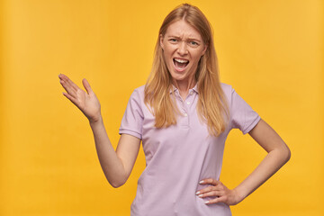 Indoor studio portrait of young ginger female with freckles raised her hand, screaming into camera with negative facial expression, isolated over yellow orange background