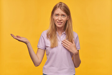 Indoor studio portrait of young ginger female with freckles looks irritated, keep her hand raised, isolated over yellow orange background