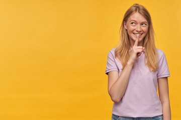 Indoor studio portrait of young ginger female with freckles showing silence gesture and smiles broadly, isolated over yellow orange background