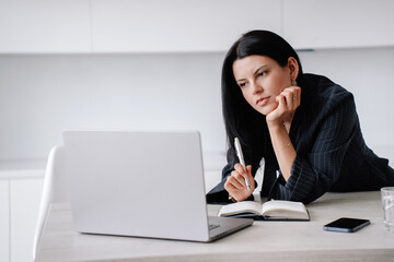 Pensive brunette tired businesswoman in suit works home sits at desk using laptop. Overloaded hispanic office manager unhappy  watching news. Financial people, fatigue. Entrepreneur.