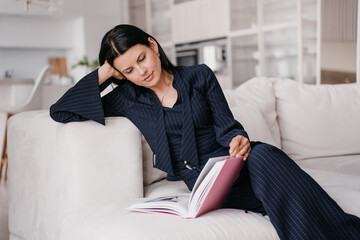 Tired caucasian businesswoman in dark blue suit sitting on couch reading book relaxing home after...