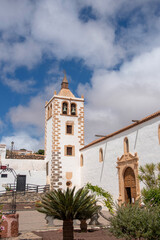 Fototapeta na wymiar Plano de detalle del campanario de piedra blanca de la iglesia en el pueblo de Betancuria en Fuerteventura, Islas Canarias, rodeado de grandes palmeras y vegetación en un día soleado.