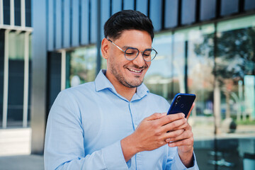 Hispanic young entrepreneur or business man smiling and using a cell phone app, wearing executive shirt and glasses. Handsome latin professional male browsing on internet with smartphone at workspace