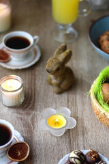 Easter eggs in the basket, bowl of cookies, chocolate pralines, Easter bunny figurine, cups of tea, glasses of juice, flowers and lit candles on the table. Selective focus.