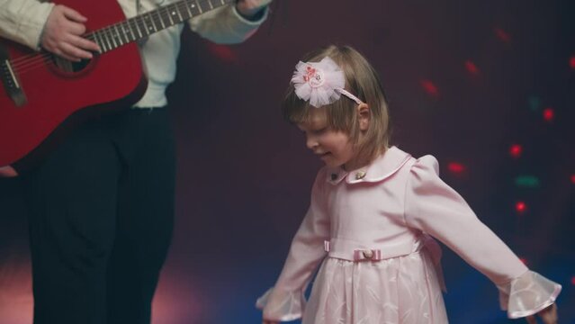 Little girl on stage in pink vintage dress, she dances, her father plays acoustic guitar. Color music is shining. Performance on stage. Early development of children