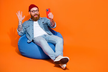 Full length photo of excited funny man wear jeans shirt sitting bean bag talking device empty space...