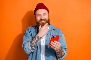 Photo of satisfied funky young man touch chin red hair beard enjoy using his phone interface download music app isolated on orange color background