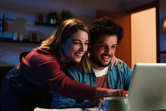 Young happy caucasian couple browsing on internet using a laptop to search sale at home. Excited husband and cheerful wife smiling and looking for new apartment sitting on a desk at living room. High