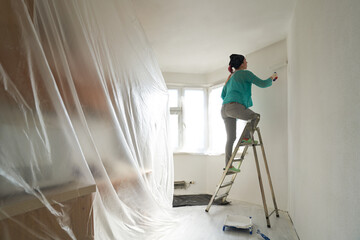 Woman stands on the stairs and paints the wall with a roller - obrazy, fototapety, plakaty