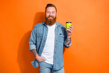 Photo of cheerful satisfied young hipster man tattoo hands hold plastic cup fresh cappuccino drinking energy isolated on orange color background