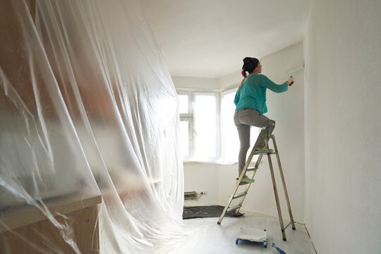Woman stands on the stairs and paints the wall with a roller