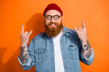 Photo of optimistic guy wear glasses red beanie cap show fingers punk symbol music lover stylish denim look isolated on orange color background