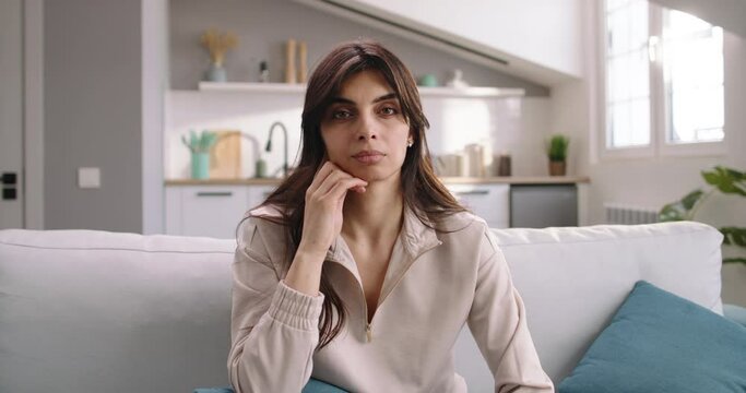 Profile of a woman who sits on a white sofa with blue pillows and carefully stares into the camera while listening to speech. POV face-to-face dialogue with woman sitting on white sofa during the day.