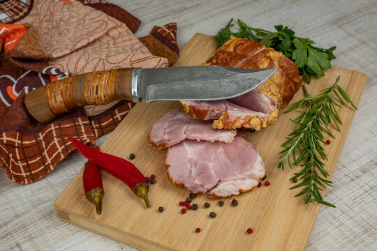 On a wooden cutting board, sliced juicy pork ham, carving knife, red hot peppers, multi-colored allspice, parsley, dill and basil. The concept of delicious pork products