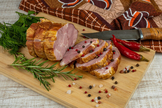 On a wooden cutting board, sliced juicy pork ham, carving knife, red hot peppers, multi-colored allspice, parsley, dill and basil. The concept of delicious pork products