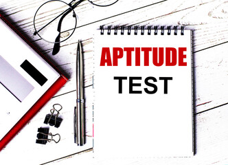 Notepad with text APTITUDE TEST, glasses, paper clips, white calculator and pen on desktop, top view.